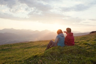 Ein Paar sitzt waehrend einer Wanderung auf der Wiese und blickt ueber das Tal.